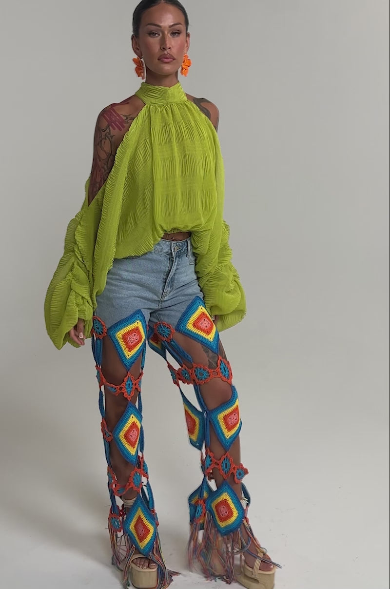 Video of woman modeling the NOT PLAYING BLOUSE in lime green with balloon sleeves and a high neck, paired with colorful open-patterned crochet pants and bold orange floral earrings, posing against a light background.