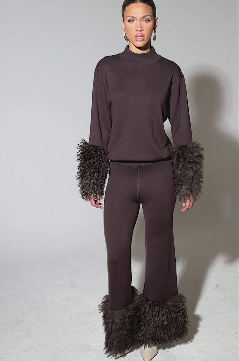 Video of woman modeling the COZY VIBES SWEATER PANT WITH FUR CUFF IN BROWN, featuring shaggy faux fur cuffs on the pant hems, paired with white heeled boots against a plain light gray background.