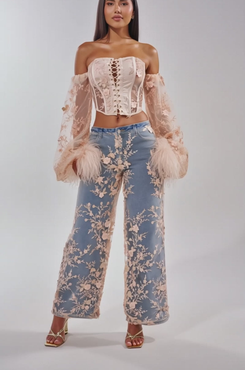A video of a woman in the FLY AWAY WITH ME FLORAL DENIM—high-rise, wide-leg jeans with a floral mesh overlay—styled with a sheer off-the-shoulder lace-up corset top and strappy heels.