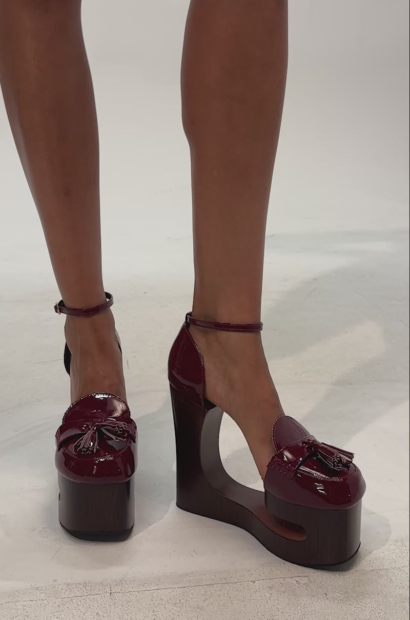 Video of woman modeling the AKIRA GOLD BIXBY BURGUNDY EMPTY WEDGE PUMP featuring a single high-heeled wedge sandal design in rich burgundy patent leather with an ankle strap, wooden platform base, and tassel detail on the toe