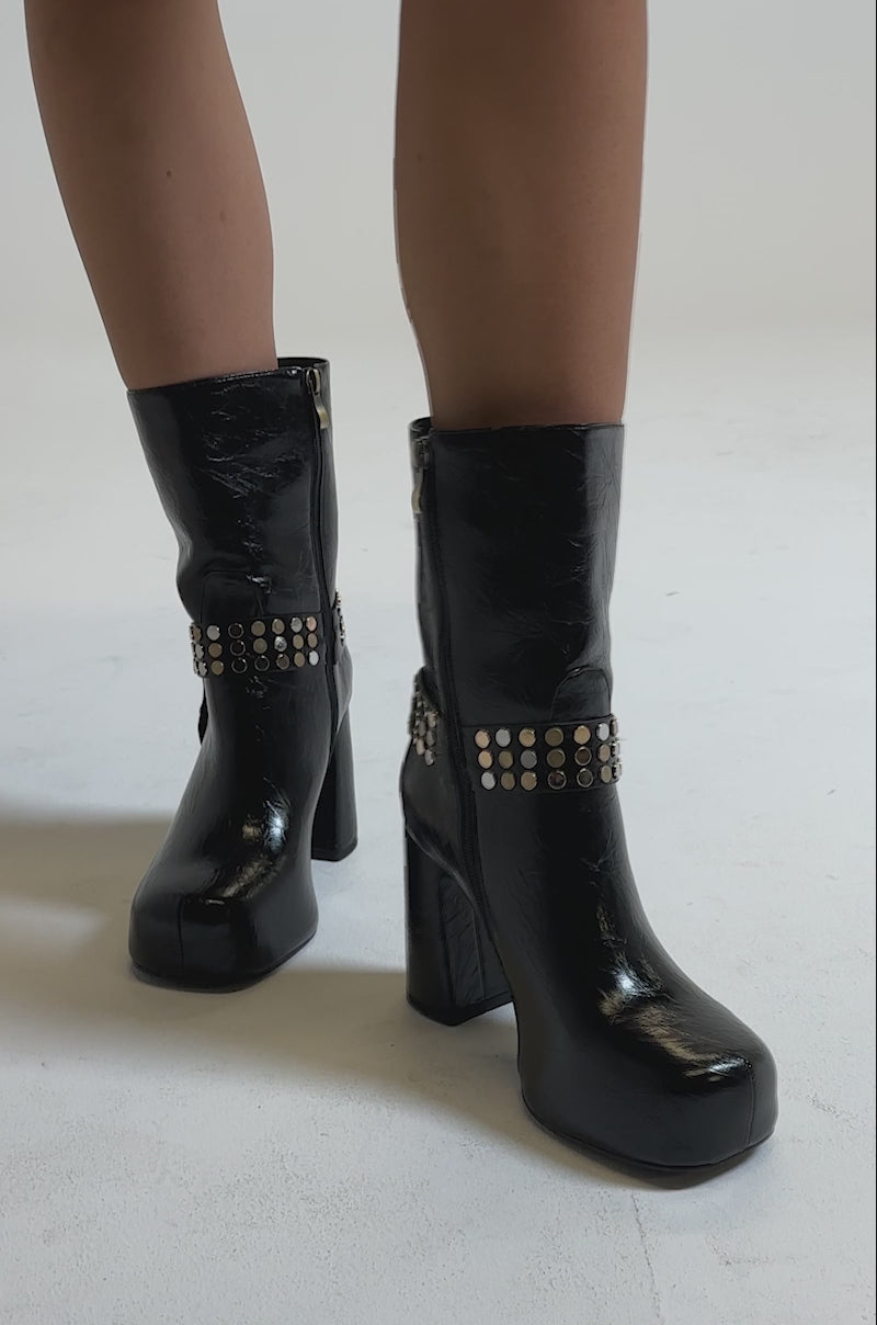 A video of a model wearing the AZALEA WANG MID CALF PU LEATHER CHUNKY BOOTIE in black with gold studs and a shiny finish, the person pairs it with a short black skirt featuring silver hooks and rings against a plain, light background.