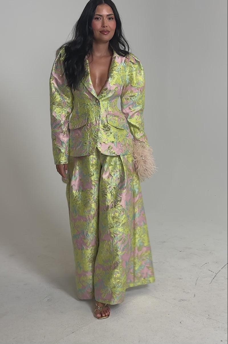 Video of woman modeling the LUCY BROCADE BLAZER and matching pants in metallic pink, green, and gold tones with a deep V-neck. She holds a light-colored feathered accessory and poses confidently.
