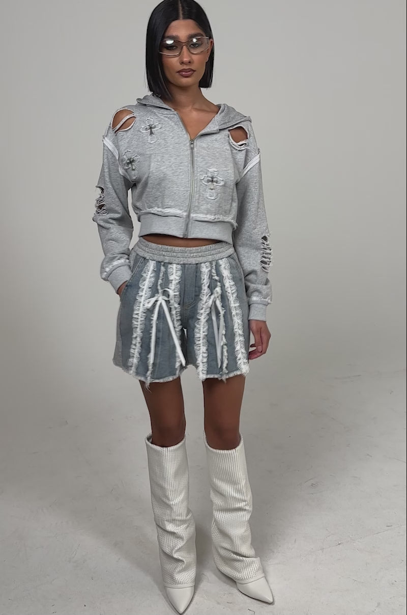 A video of a woman wearing the TIED AND LACED DENIM SHORTS with lace ruffle and tie details, a light gray distressed cropped hoodie, oversized white textured boots, and small sunglasses.
