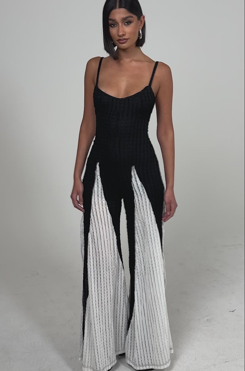 A video of a woman in  the ALL DAY EVERYDAY COLOR BLOCK WOVEN JUMPSUIT—a sleeveless black and white wide-leg style—against a plain light background.