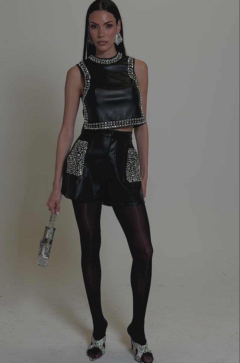 A video of a woman in the DANCE THE NIGHT AWAY FAUX LEATHER SHORTS with rhinestone embellishments, a black sleeveless crop top, sheer tights, heeled sandals, and a metallic handbag against a plain neutral background.