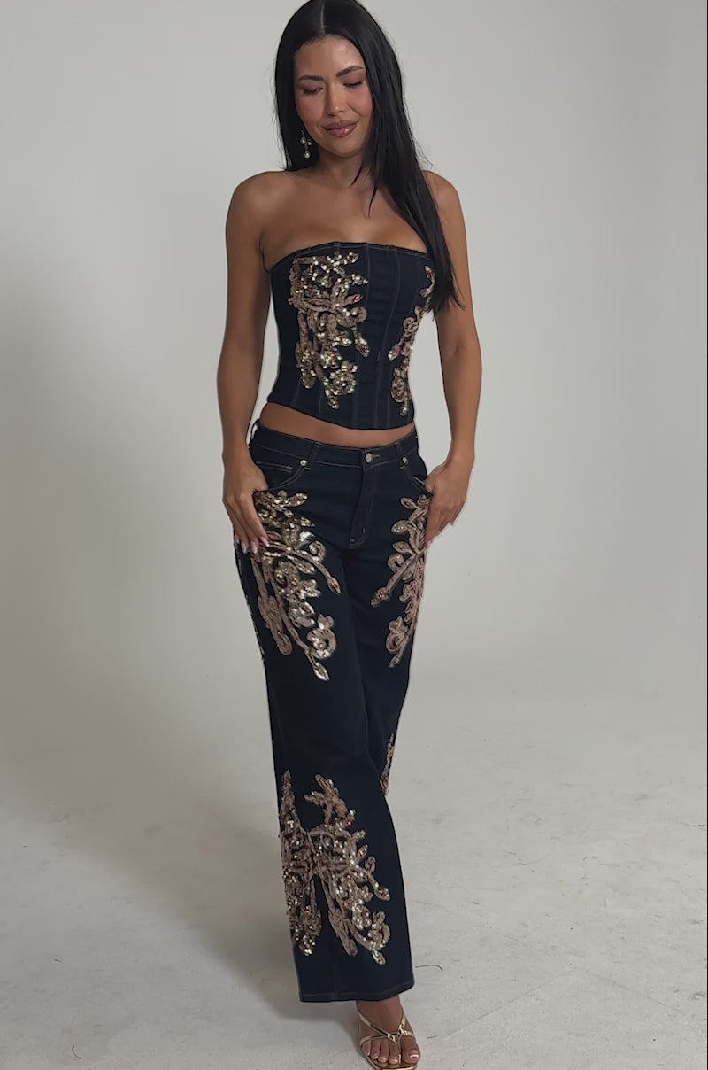 Video of woman modeling the MALIBU EMBELLISHED WIDE LEG DENIM—a navy blue strapless corset top paired with matching wide-leg pants adorned with intricate gold embroidery—and gold heeled sandals.