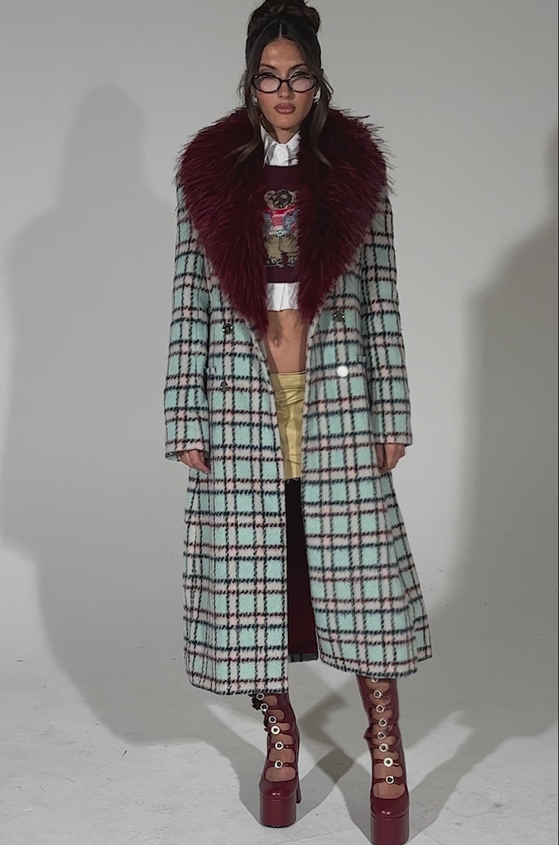 A video of a woman posing against a plain light background wearing the TEA FOR TWO FUR TRIM TRENCH IN BLUE, styled with glasses, tan shorts, tall red lace-up platform boots, and her hair up.