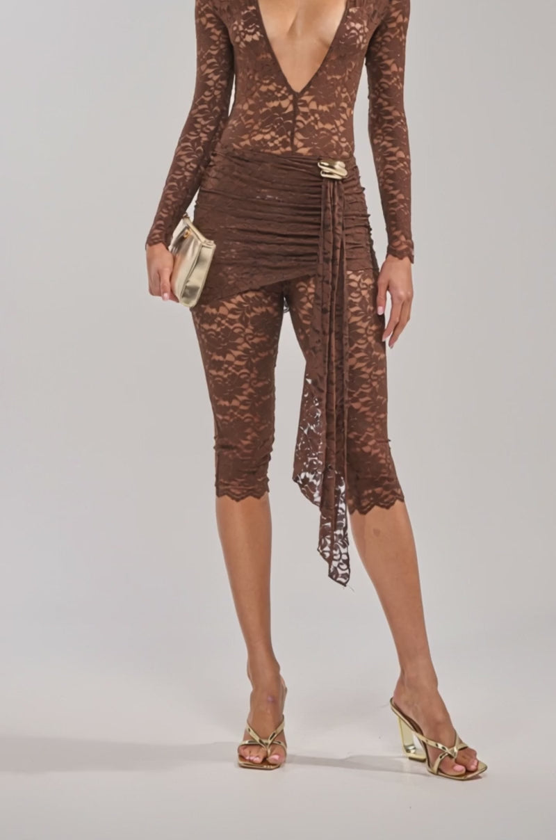 Video of woman modeling the ICONIC BEHAVIOR LACE CAPRI JUMPSUIT, a sheer brown lace jumpsuit with a deep V neckline and 3/4 length pants. She completes her night-out look with a gold clutch, gold earrings, curly hair, and gold high-heeled sandals.