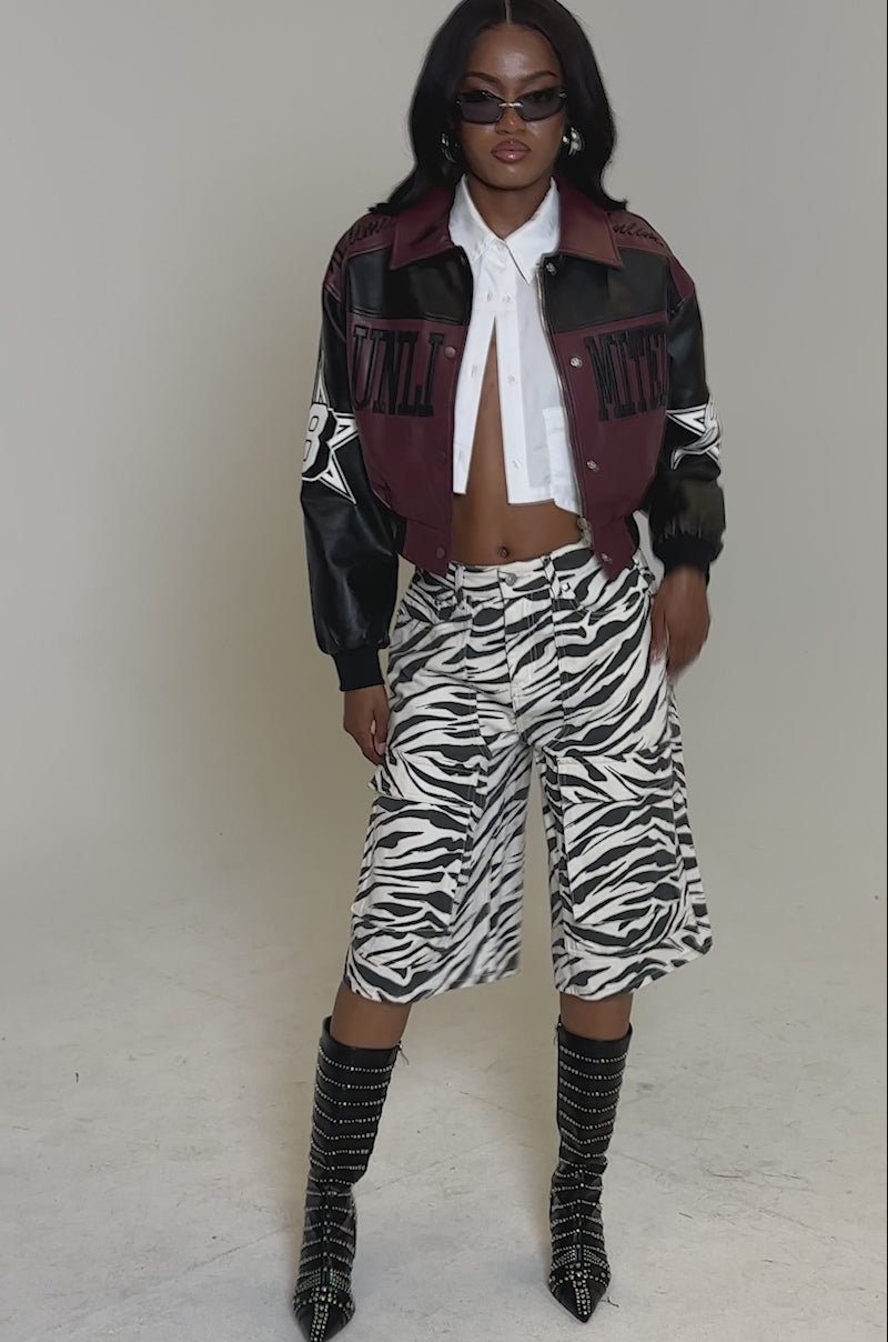 A video of a person posing in the HARD TO FORGET MOTO BOMBER, paired with a white cropped shirt, zebra-print knee-length shorts, black studded boots, and sunglasses against a plain background.