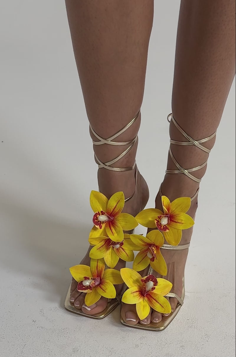 A video of a model wearing the AZALEA WANG CRESCENDO Gold Stiletto Sandal, featuring metallic gold heels adorned with large, vibrant yellow orchid flowers.