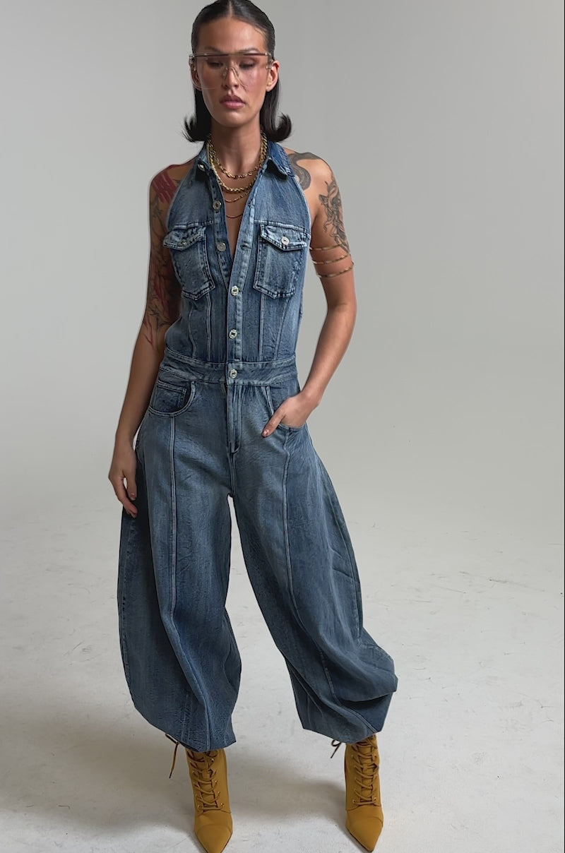 A video of a person wearing the JETSETTER JUMPSUIT IN MEDIUM BLUE DENIM with wide legs, yellow lace-up boots, clear glasses, and layered necklaces.