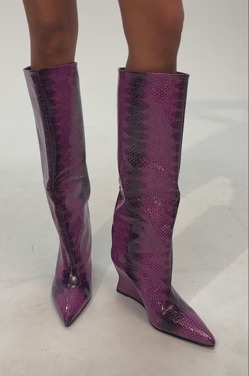 Video of woman modeling the AZALEA WANG BRUN PURPLE SNAKE WEDGE BOOT, which features knee-high, pointed-toe wedges with a shiny purple and black snakeskin pattern