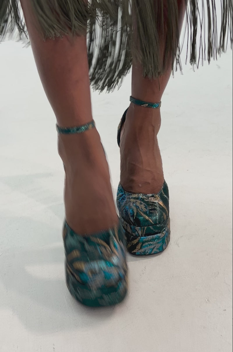 Video of woman modeling the AZALEA WANG ZIPPHORA BLUE BROCADE CHUNKY PUMP heels with ankle straps stands on a patterned blue and gold carpet.