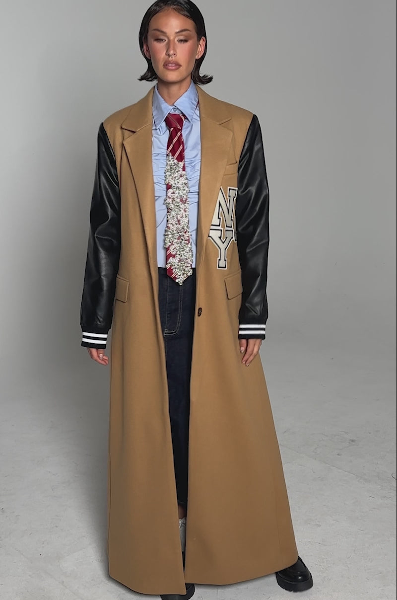 A video of a model wearing the BRENTWOOD 2.0 VARSITY TRENCH with faux leather sleeves, a blue shirt, red striped tie, dark wide-leg jeans, and chunky black shoes, this look blends formal and casual styles with confidence.