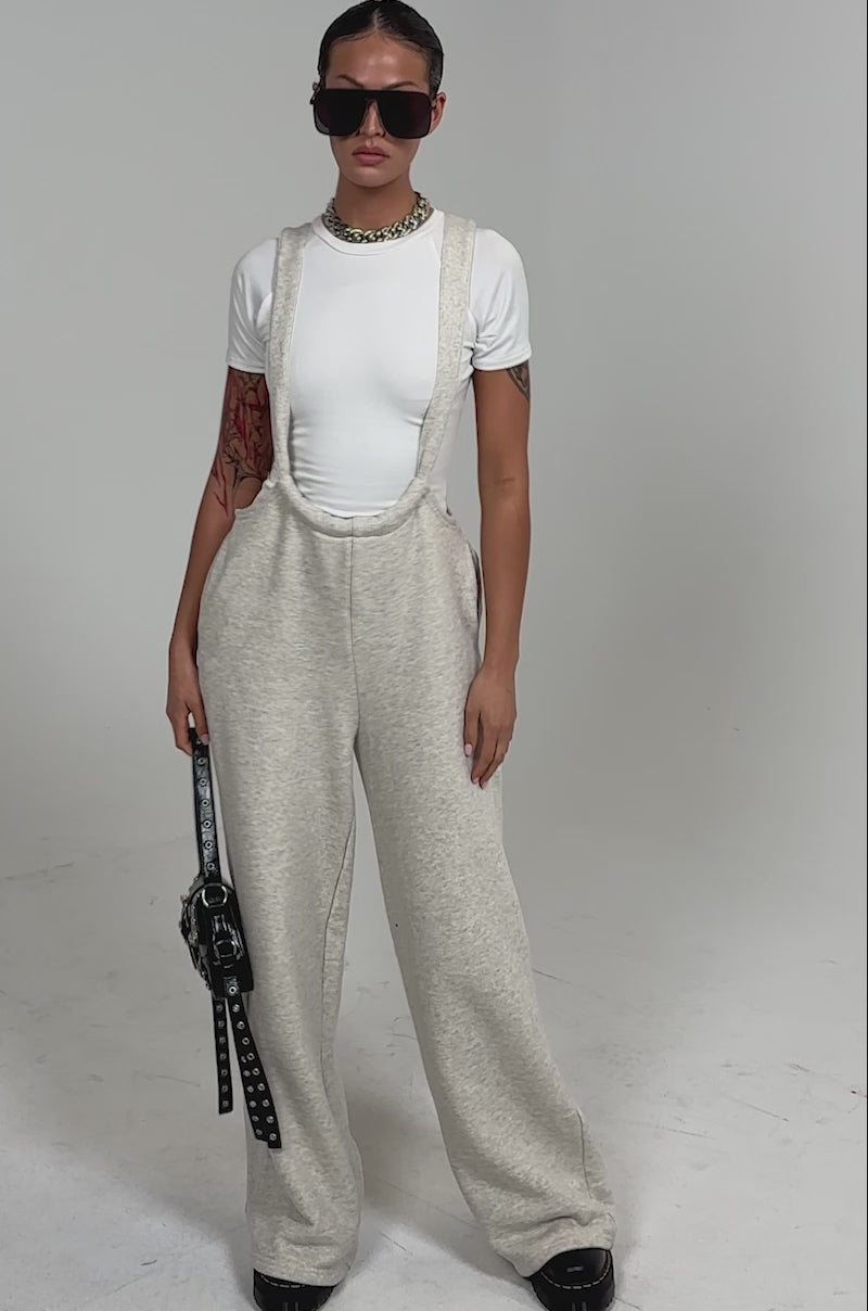Video of woman modeling the ALESSANDRA OVERALLS in beige with a fitted white tee, chunky black shoes, sunglasses, a silver chain necklace, and holding a black handbag