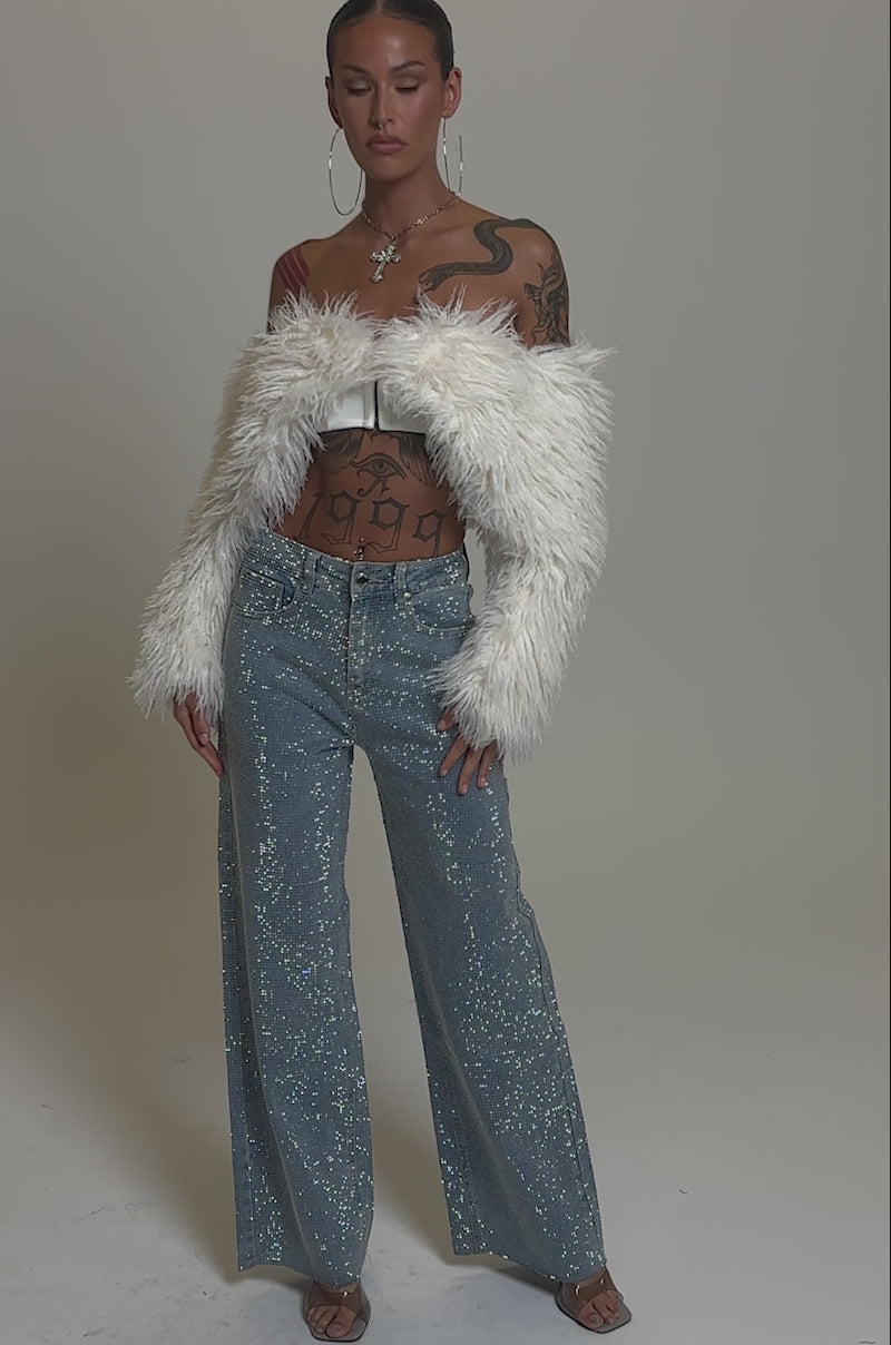 Video of woman modeling DANCING QUEEN RELAXED DENIM high-waisted wide-leg jeans, a cream faux fur shrug, and clear heels. She accessorizes with hoop earrings, layered necklaces, and reveals tattoos on her torso and neck against a gray backdrop.