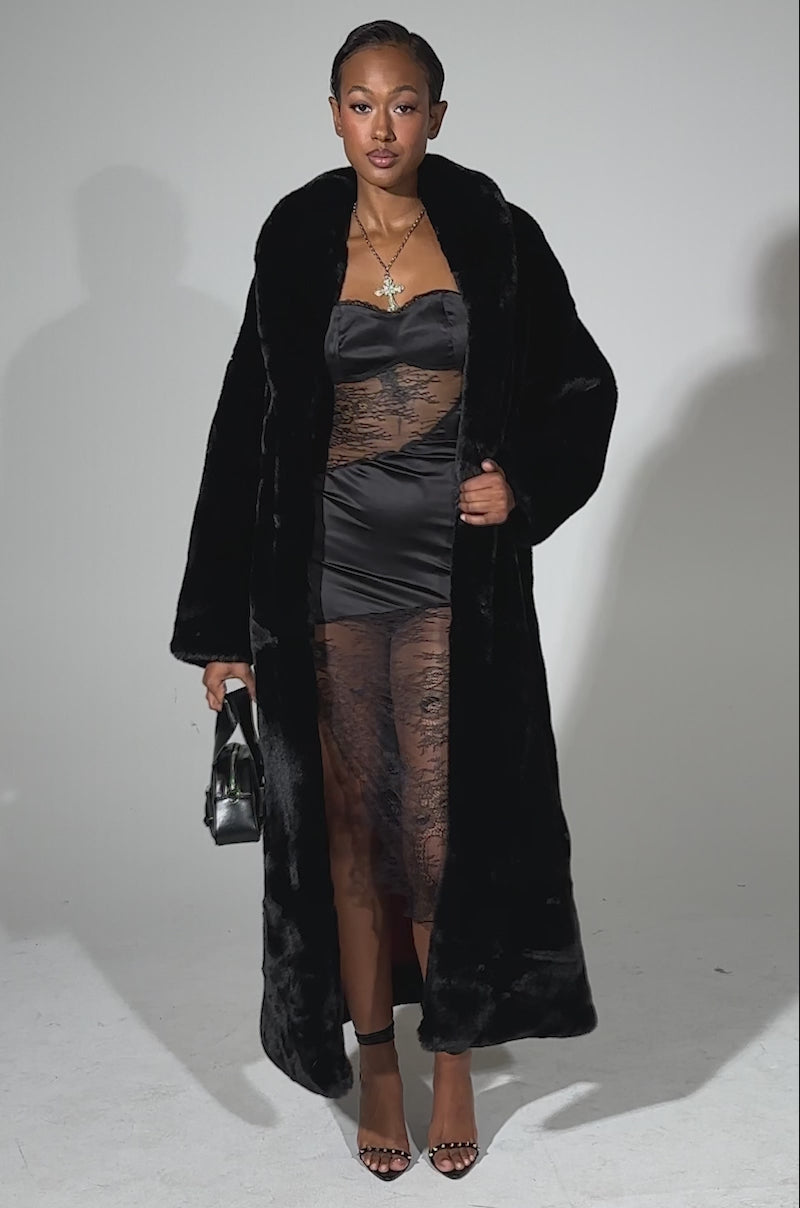 A video of a woman posing confidently against a light background in the DAYANNE LONG SATIN LINING FAUX FUR TRENCH COAT over a sheer lace black dress with a high slit, styled with strappy heels, layered necklaces, and earrings.