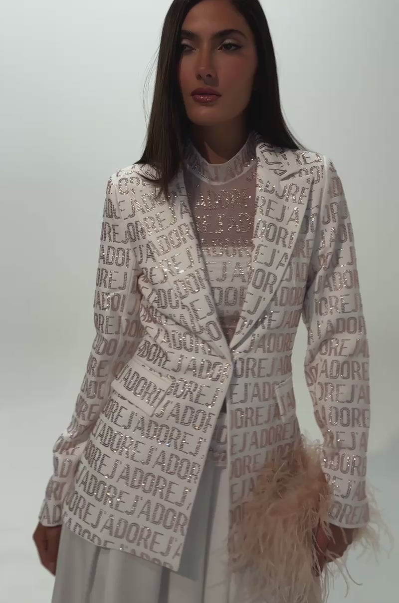 A video of a woman with straight brown hair in the J'ADORE EMBELLISHED BLAZER IN WHITE over a sheer high-neck blouse and white skirt, holding a feathery light beige accessory in one hand.