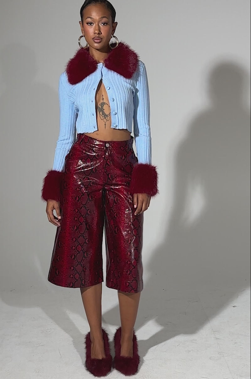 A video of a woman standing confidently against a plain light background, wearing the EXTRA IS MY MIDDLE NAME SNAKE BERMUDA SHORT styled with a light blue cropped sweater, red fur trim, red fluffy heels, gold hoop earrings, and showing a tattoo on her chest.