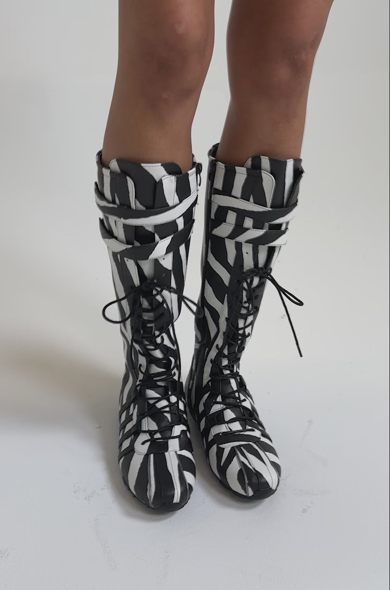 A video of a model wearing the OZZY ZEBRA FLAT SNEAKER boots, featuring a striking black and white zebra stripe pattern.