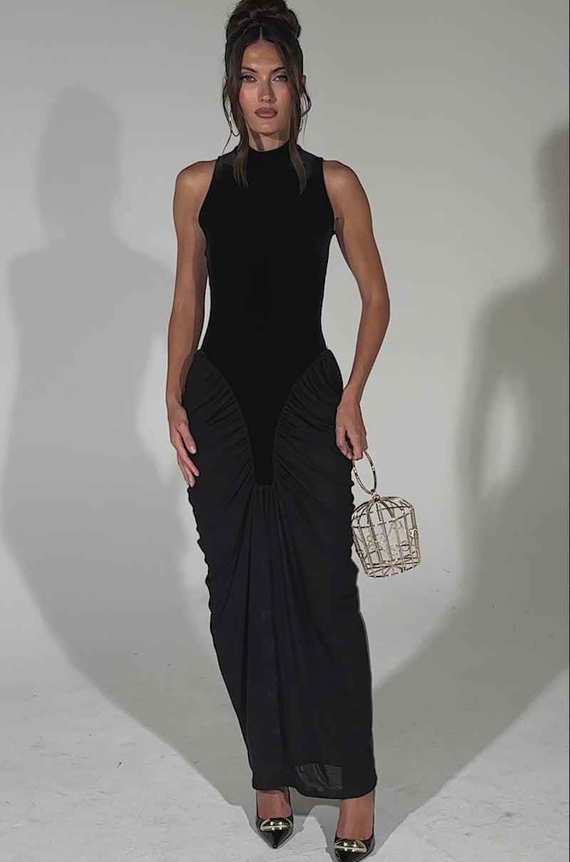 A video of a woman wearing the OUT FOR THE NIGHT MOCK NECK MAXI DRESS in black with ruched detailing, stands against a plain background, holding a gold cage-shaped bag and wearing black pointed-toe heels with gold accents.
