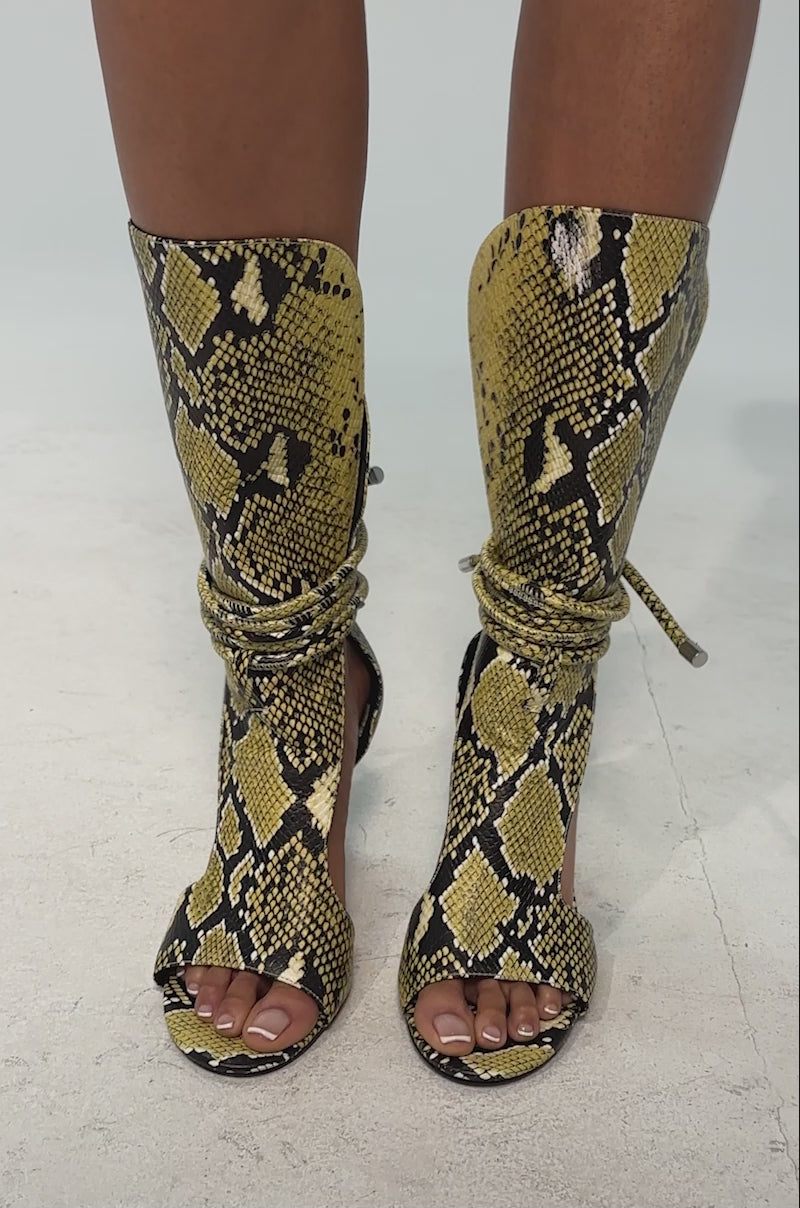 A video of a model wearing the AZALEA WANG BOCKER YELLOW SNAKE STILETTO LACE UP TALL SHAFT SANDAL featuring yellow and black faux snakeskin, an open toe, thin high heel, tall shaft, and a wraparound lace-up front with drawstring ankle closure.