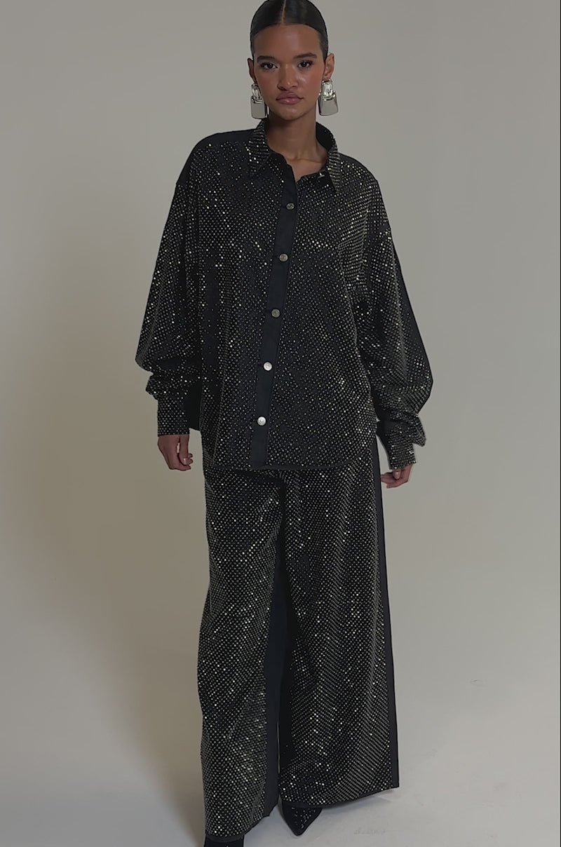 Video of woman modeling the BLING BLING DENIM BUTTON DOWN TOP IN BLACK with rhinestones, wide-leg pants, and large silver geometric earrings, standing against a plain grey background.