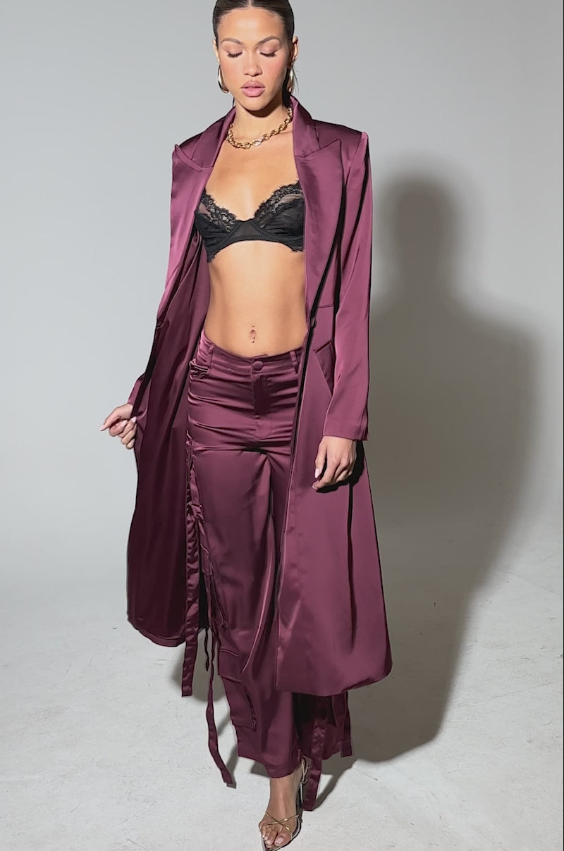 Video of woman modeling the TAKE ME EVERYWHERE SATIN CARGO PANT, featuring a high-rise waist, wide legs, maroon satin fabric, and ribbon-like side straps, paired with clear high-heeled sandals against a plain light background