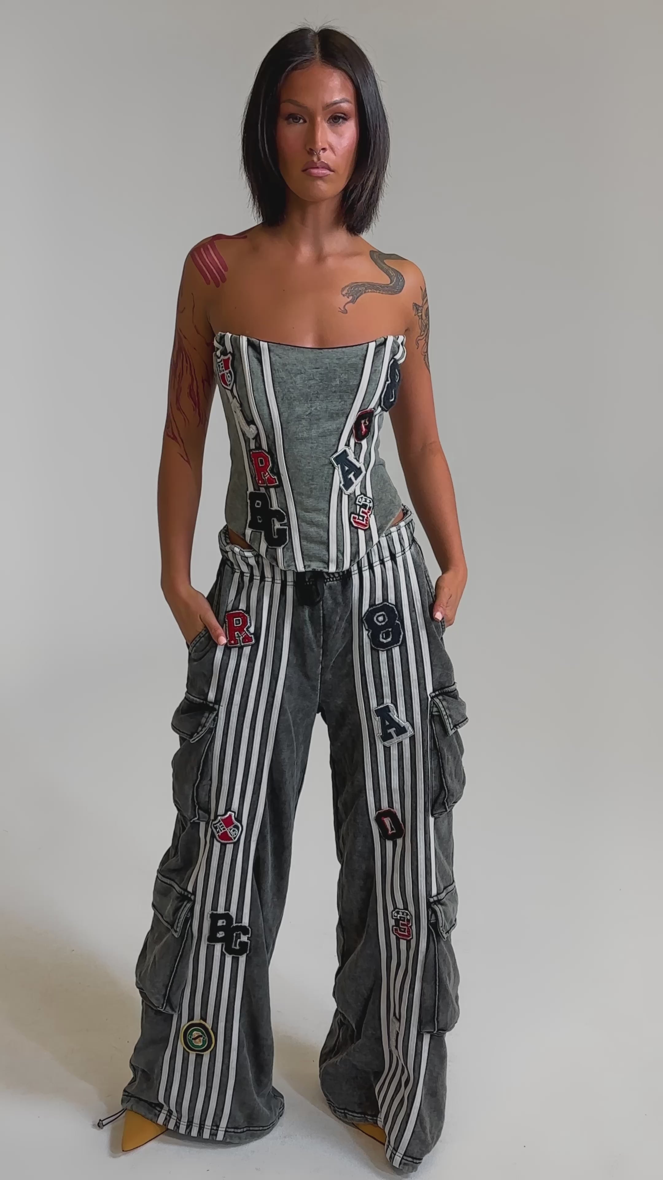 A video of a woman wearing the ROCK AND ROLL CORSET, paired with gray striped pants featuring ruffled sides and colorful letter patches. 