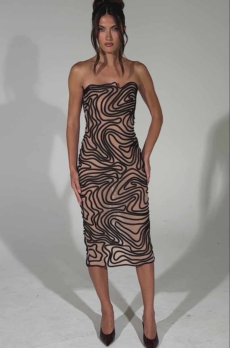A video of a woman posing on a plain light background wearing the OUT OF THIS WORLD MIDI DRESS, featuring a strapless neckline, abstract ribbed detailing, bold black swirling patterns, and paired with black pointed heels.