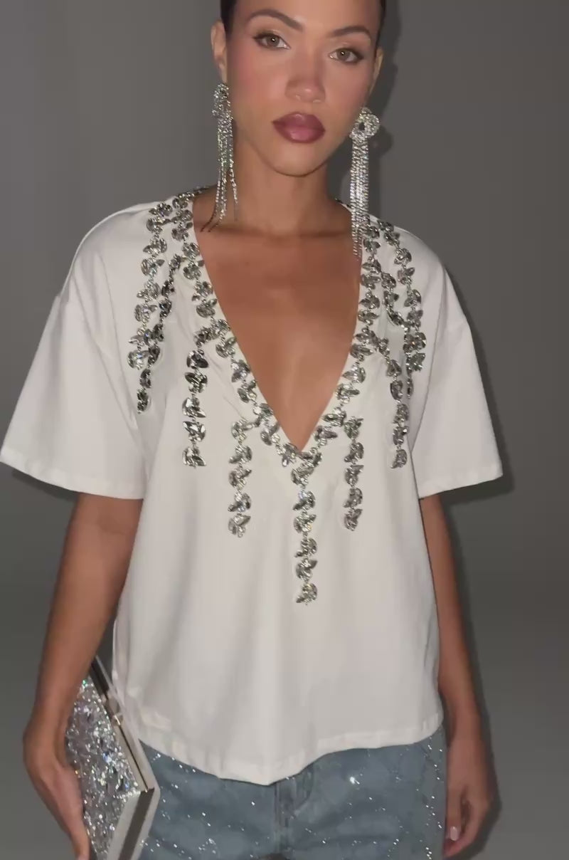 Video of woman modeling the TOO DEEP RHINESTONE TEE, light blue jeweled jeans, long rhinestone earrings, and holding a silver clutch.