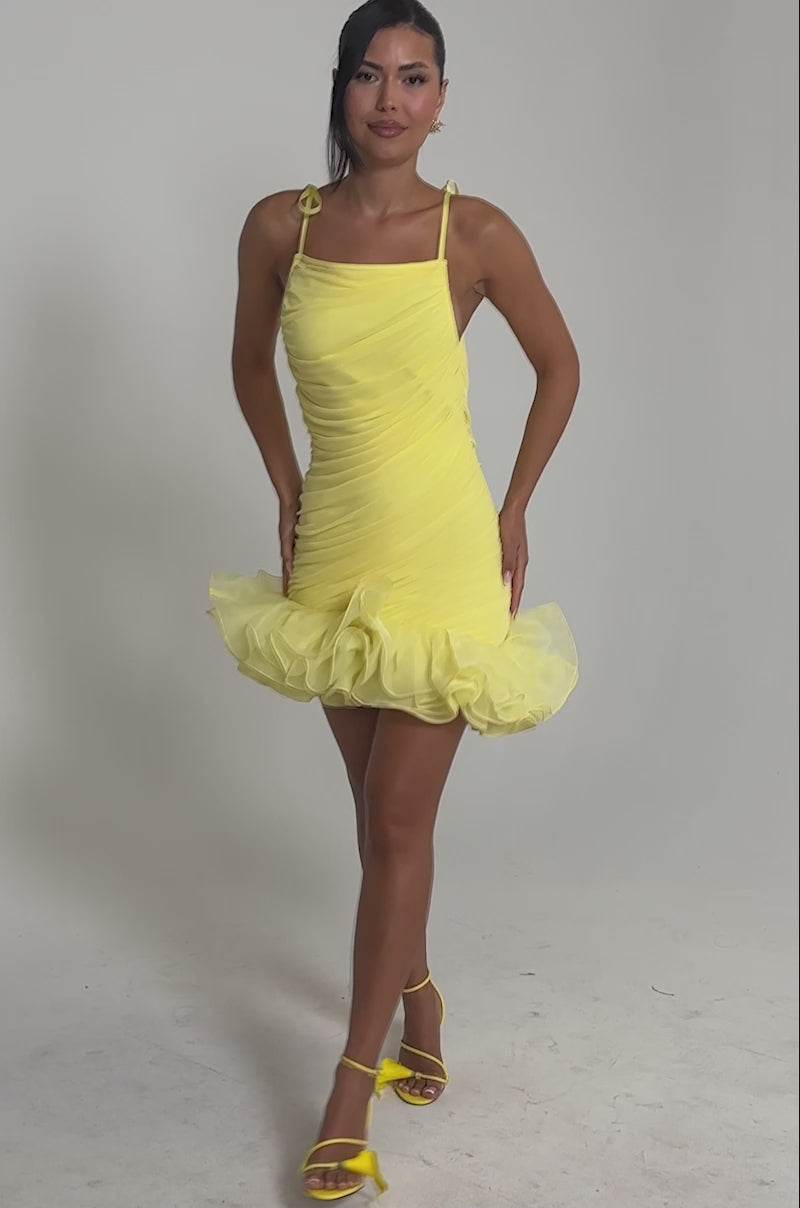 Video of woman modeling the PRINCESS PEPLUM MINI DRESS IN YELLOW with ruffled chiffon and tulle details. Her dark hair is styled up, and she poses against a plain light background.