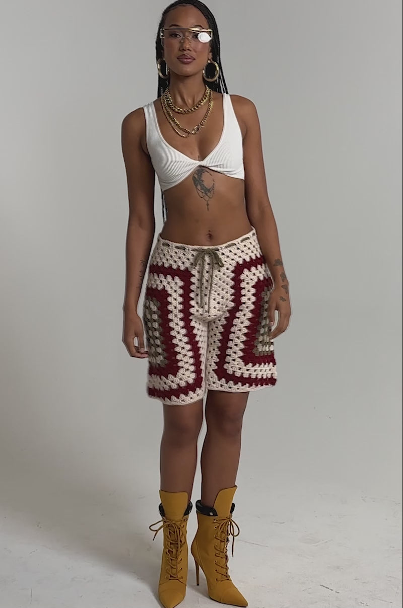 A video of a woman in the VACATION VIBES CROCHET SHORT, paired with a white bralette, yellow lace-up boots, gold jewelry, and clear glasses.
