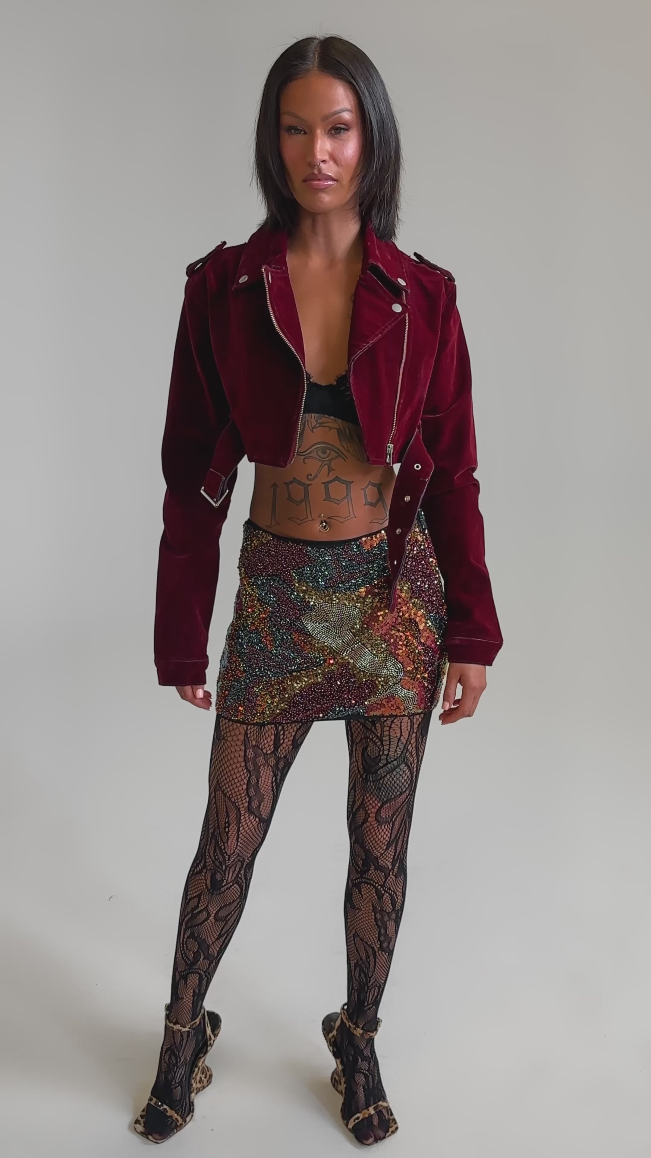 A video of a woman wearing the LULU VELVET DENIM MOTO jacket, a black lace bra, multicolored sequin mini skirt, patterned lace tights, and high heels.