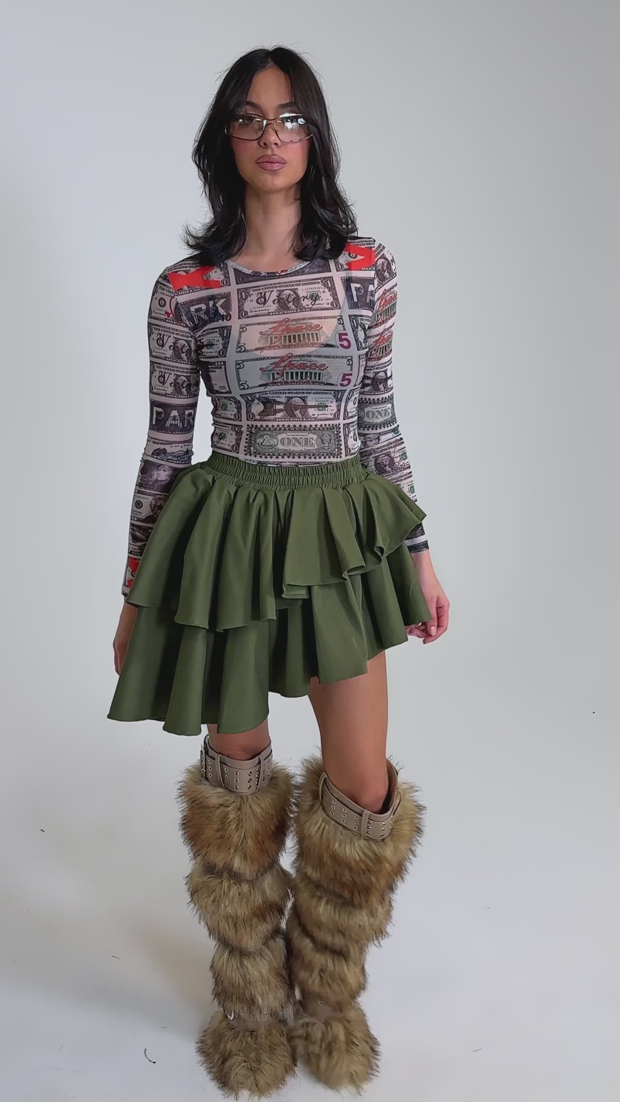 A video of a woman in sunglasses, a colorful graphic long-sleeve top, the OLIVIA OLIVE MINI PLEATED SKIRT, and tall furry brown boots standing against a plain gray background.