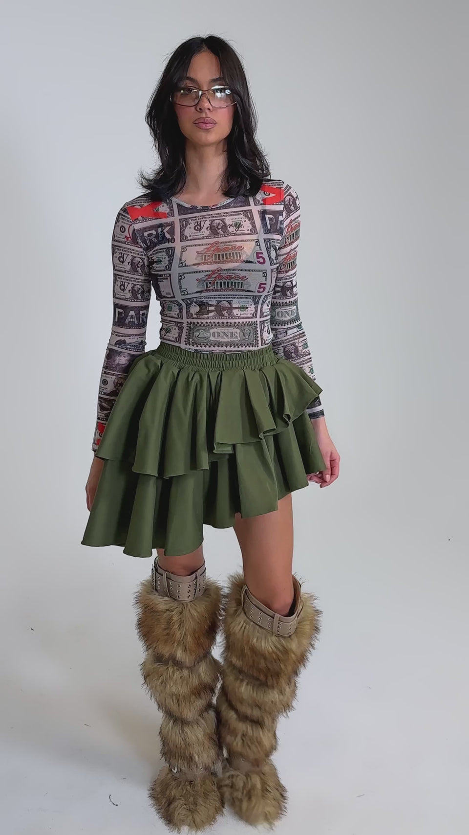 A video of a woman in sunglasses, a colorful graphic long-sleeve top, the OLIVIA OLIVE MINI PLEATED SKIRT, and tall furry brown boots standing against a plain gray background.