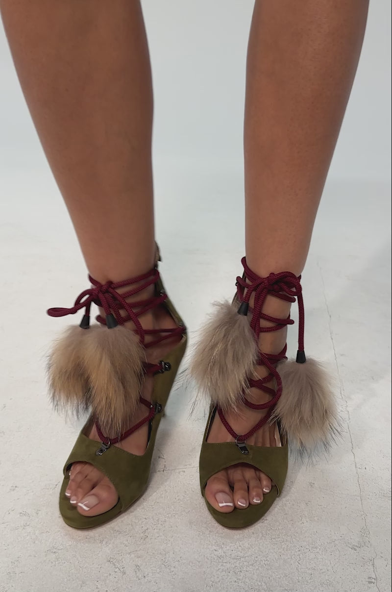 A video of a model wearing the AZALEA WANG KARLA OLIVE FAUX SUEDE FUR EMBELLISHED STILETTO SANDAL featuring olive green faux suede, a peep toe, red rope lace-up with silver eyelets, a faux fur ankle accent, and a textured stiletto heel.
