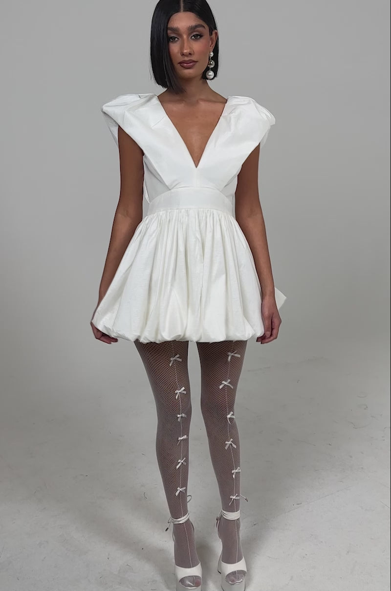 Video of woman modeling the FAR GONE WHITE MINI DRESS, a short bubble skirt style with a plunging V-neckline, paired with white fishnet tights featuring small front bows and white platform heels against a light background.