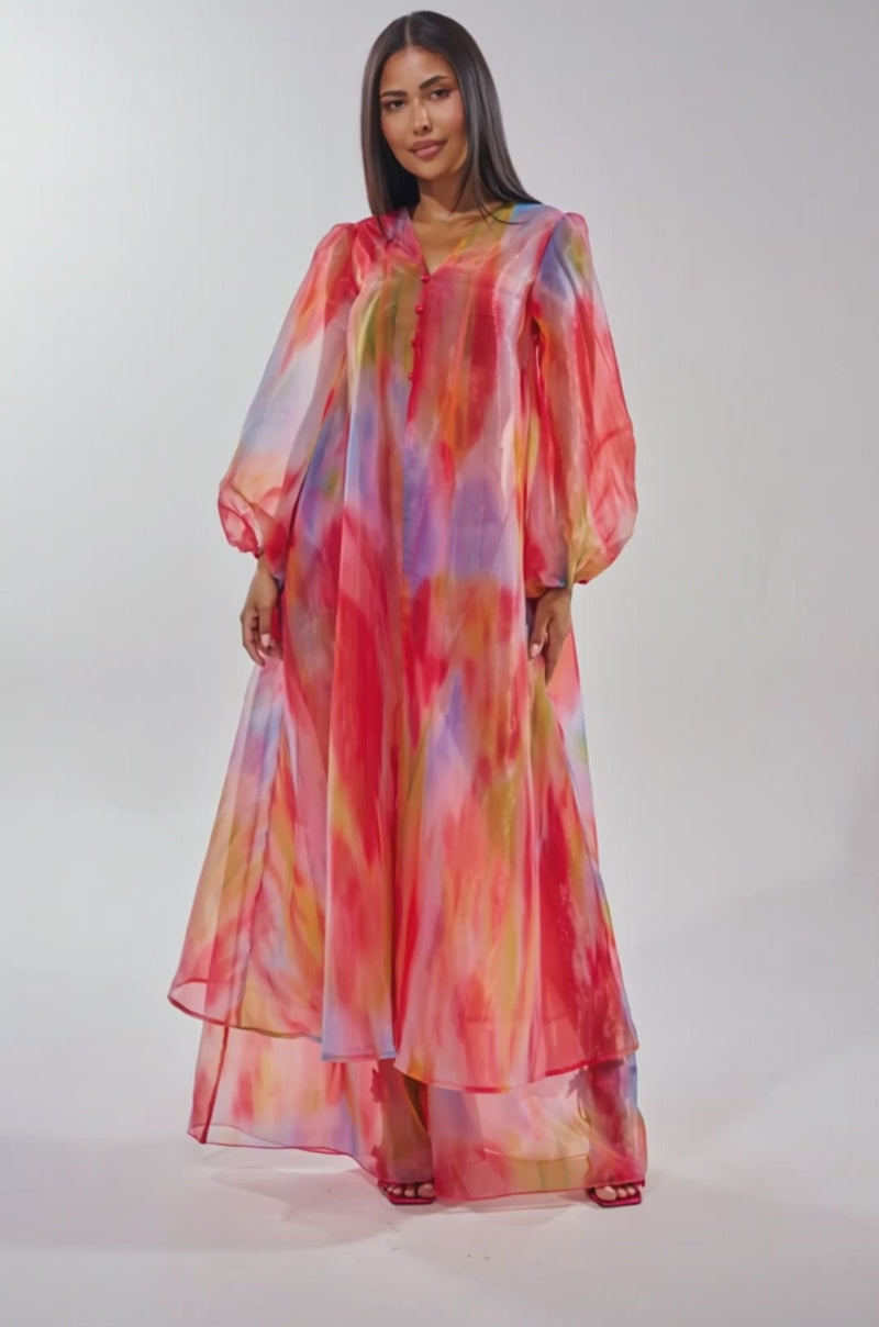 A video of a woman with straight hair wearing the INTERESTED IN YOU DUSTER, a sheer, flowing longline maxi dress featuring a colorful watercolor wash in red, blue, yellow, and pink. She completes the look with red sandals.