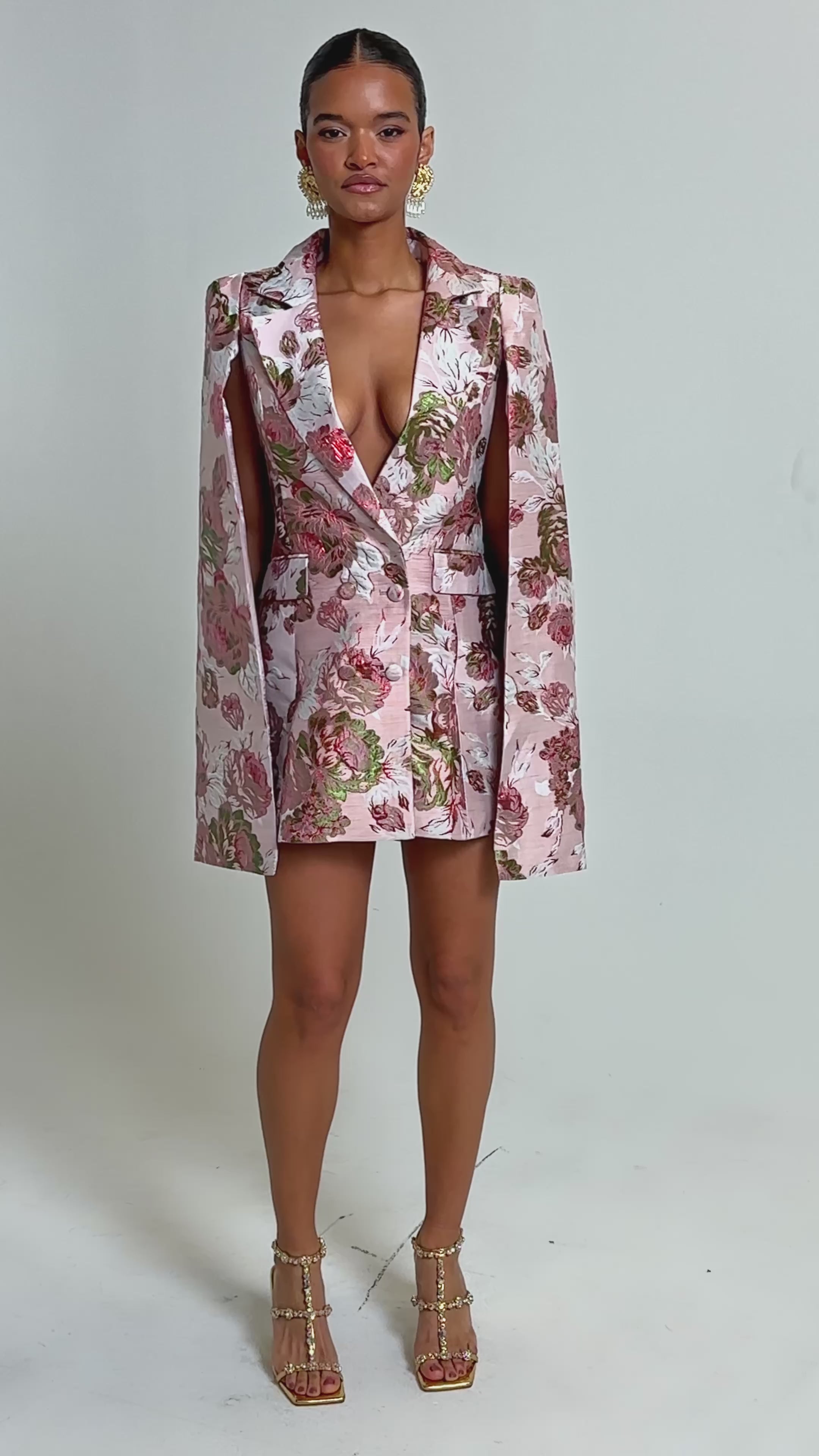 Video of woman modeling the IMANI Brocade Cape Sleeve Blazer Dress in Pink, featuring a deep V-neckline and gold statement earrings