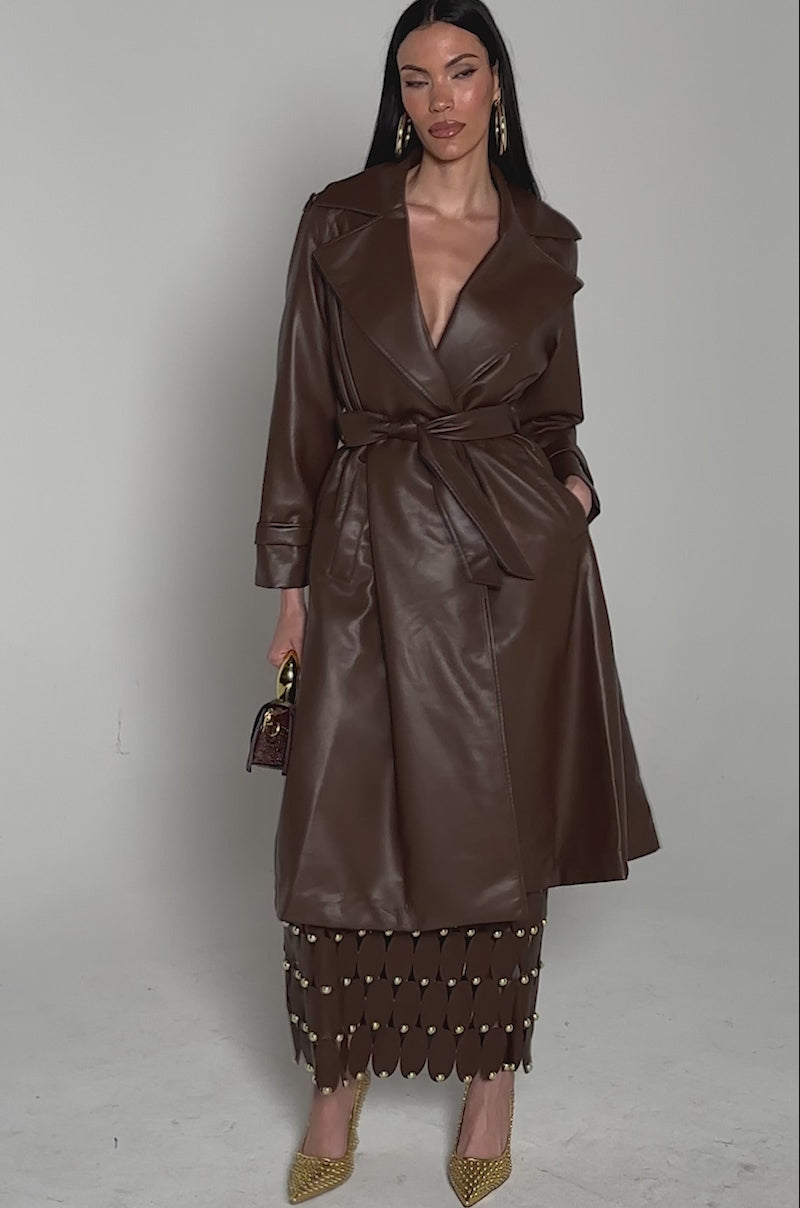 Video of woman modeling the ESPRESSO MARTINI FAUX LEATHER TRENCH over a brown skirt with gold heels, hoop earrings, gold bracelets, and a brown textured handbag, posing against a plain background.