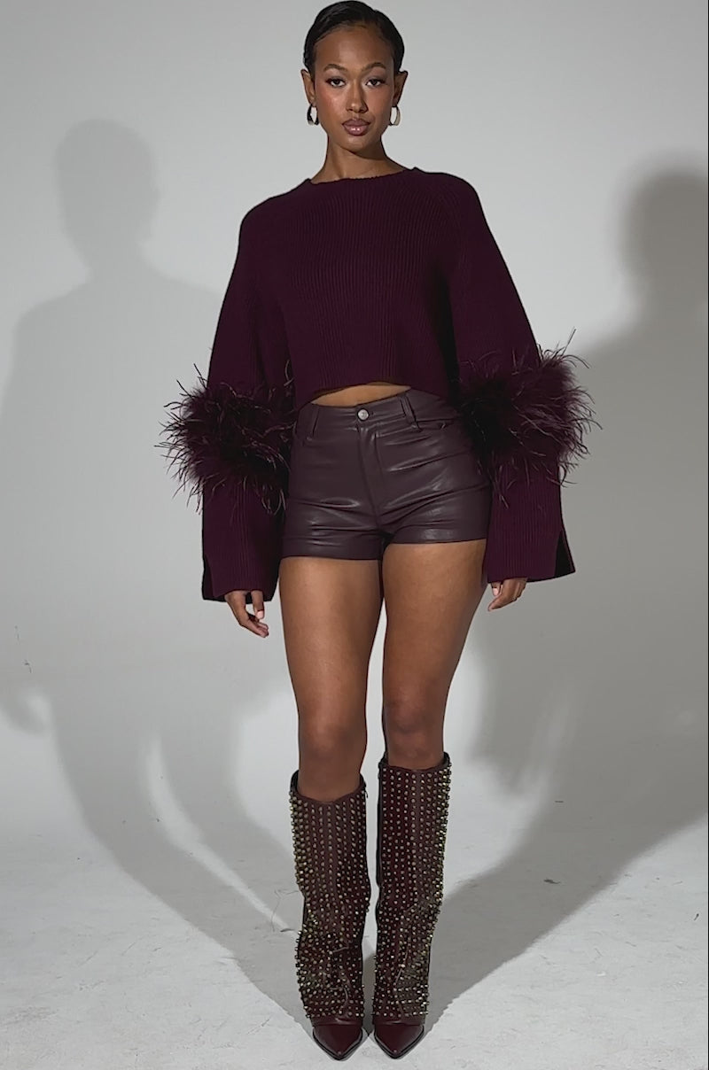 A video of a woman standing confidently in the MILLY FEATHER TRIM SWEATER with burgundy accents, paired with high-waisted leather shorts and gold-studded knee-high boots, set against a plain light background.