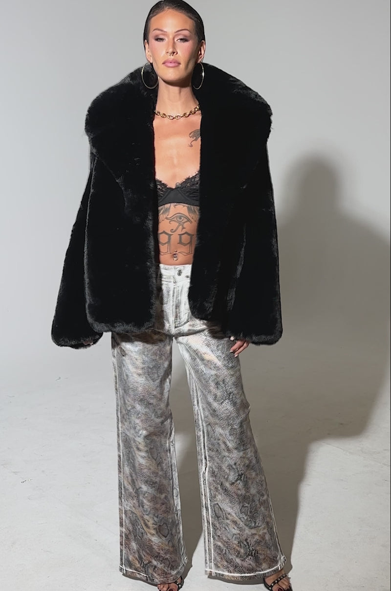 A video of a woman wearing ADDICTED TO YOU METALLIC SNAKE WIDE LEG DENIM makes a bold statement, pairing the standout jeans with a black faux fur coat, black lace bralette, and black open-toe heels against a plain background.