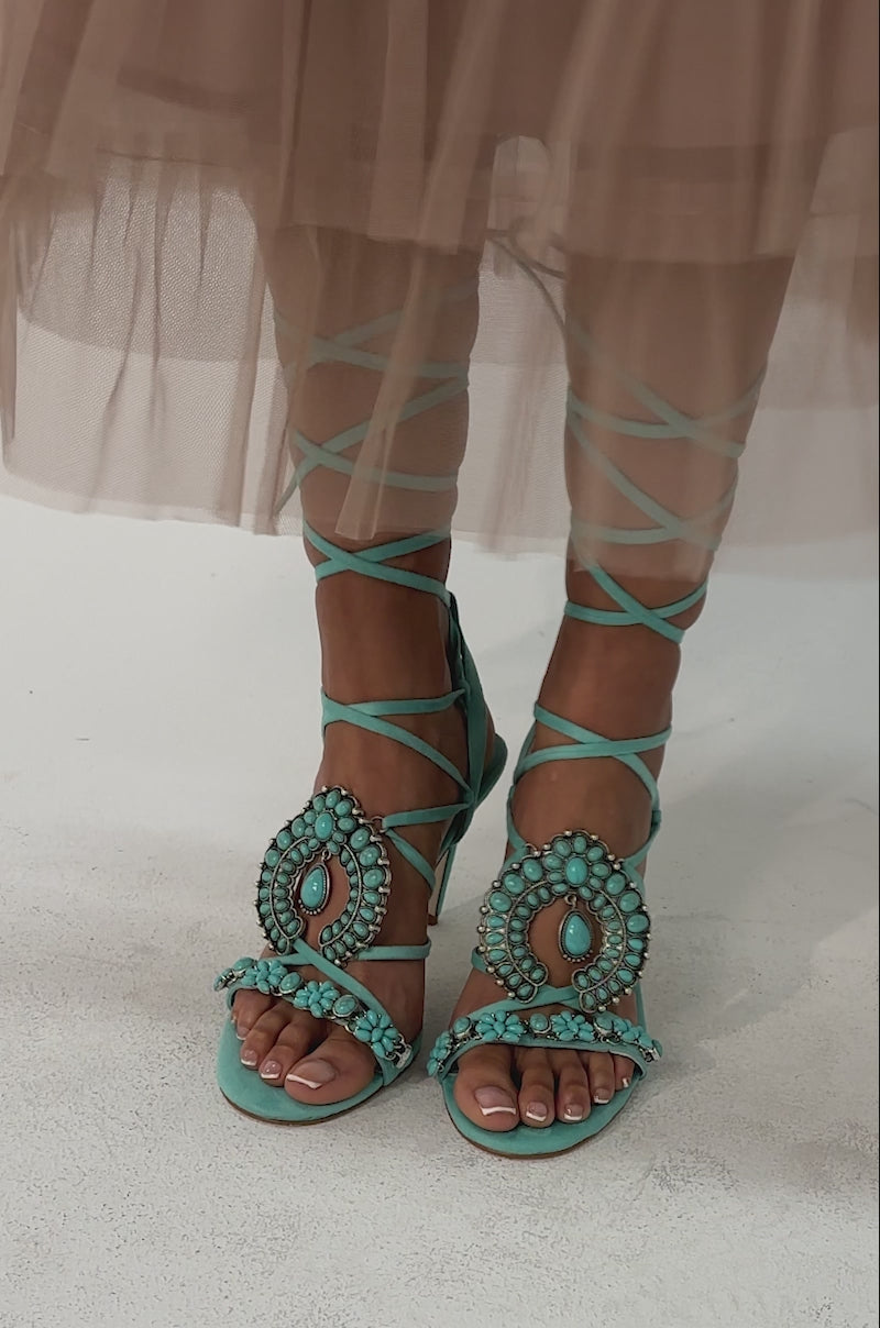 Video of woman modeling the AKIRA GOLD CLIPSE AQUA HARDWARE EMBELLISHED STILETTO SANDAL, featuring ornate beadwork and crisscrossing blue ankle straps.