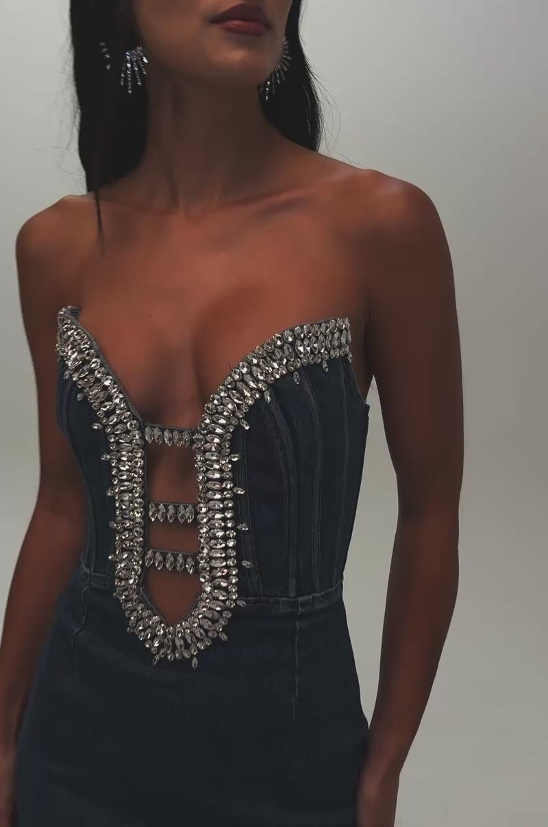 Video of a woman modeling the FEELIN THE LOVE RHINESTONE DENIM MINI DRESS, a woman poses with one hand on her hip against a plain background, showcasing the strapless silhouette adorned with rhinestones and metallic embellishments along the neckline and bodice.