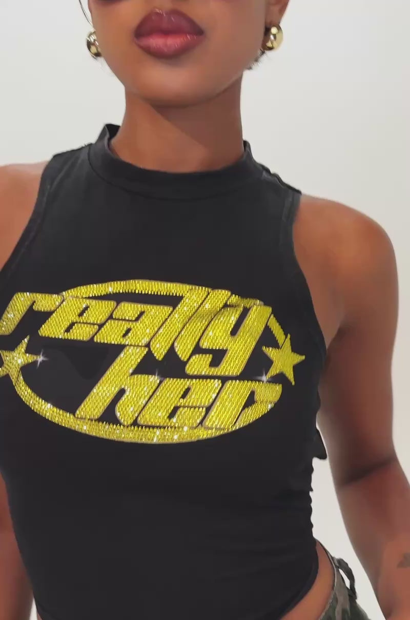 Video of woman modeling the REALLY HER TIE-BACK TANK TOP—a sleeveless black crop top with yellow text and stars—paired with large yellow sunglasses and high-waisted camo pants adorned with silver studs, this woman's confident look is the perfect festival outfit against a plain background.