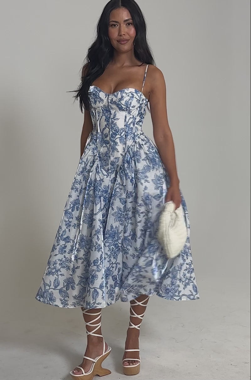 Video of woman modeling FEELING FLORAL MAXI DRESS in blue and white, featuring a fit-and-flare silhouette. She pairs it with strappy beige sandals and a white textured clutch bag, posing against a plain background.