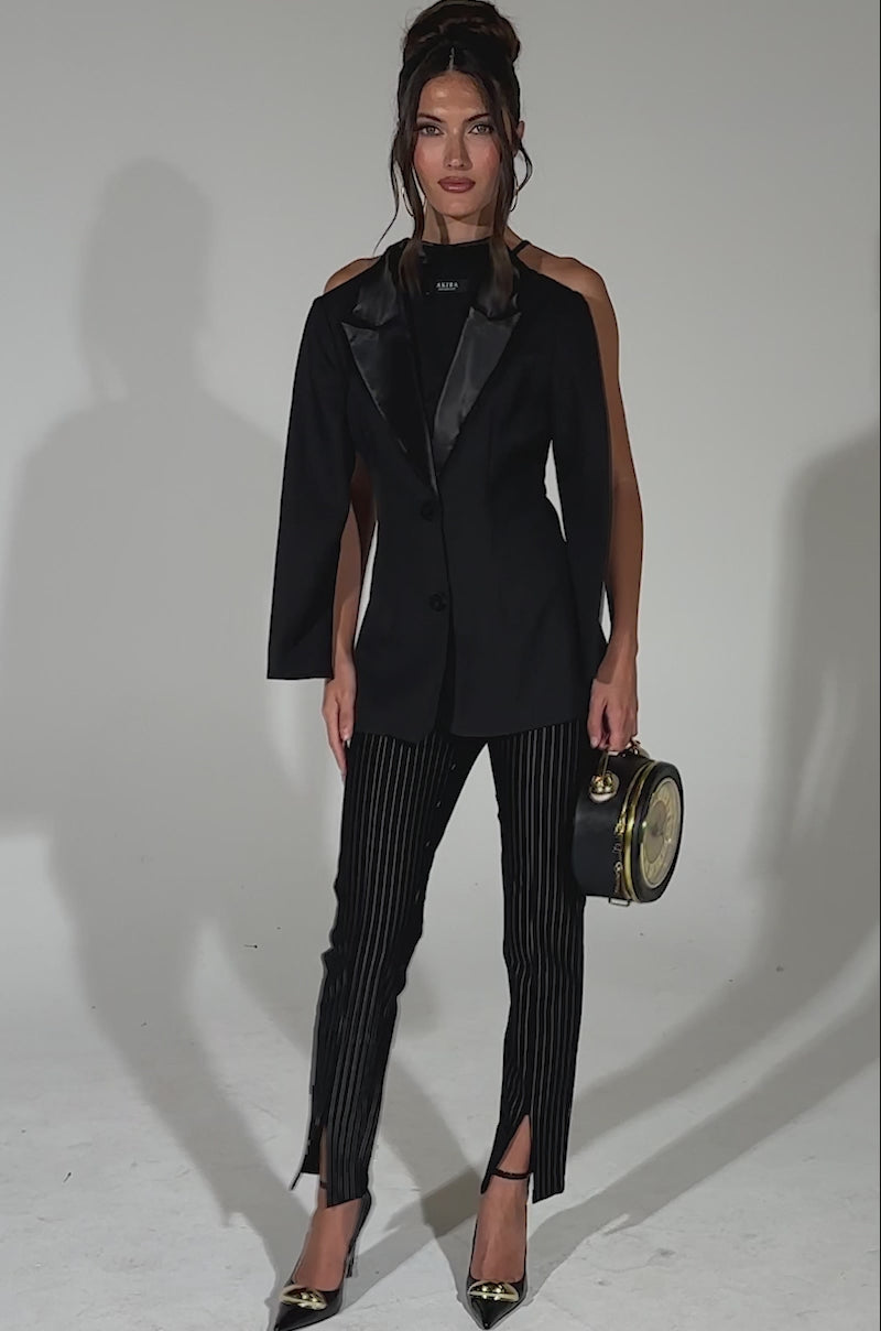 A video of a woman posing against a light background wearing the TELL ME ABOUT IT BUTTON DOWN BLAZER BLOUSE with one sleeve, pinstripe suiting fabric pants with front slits, black heels, and a round clock-inspired handbag; her hair is styled up.