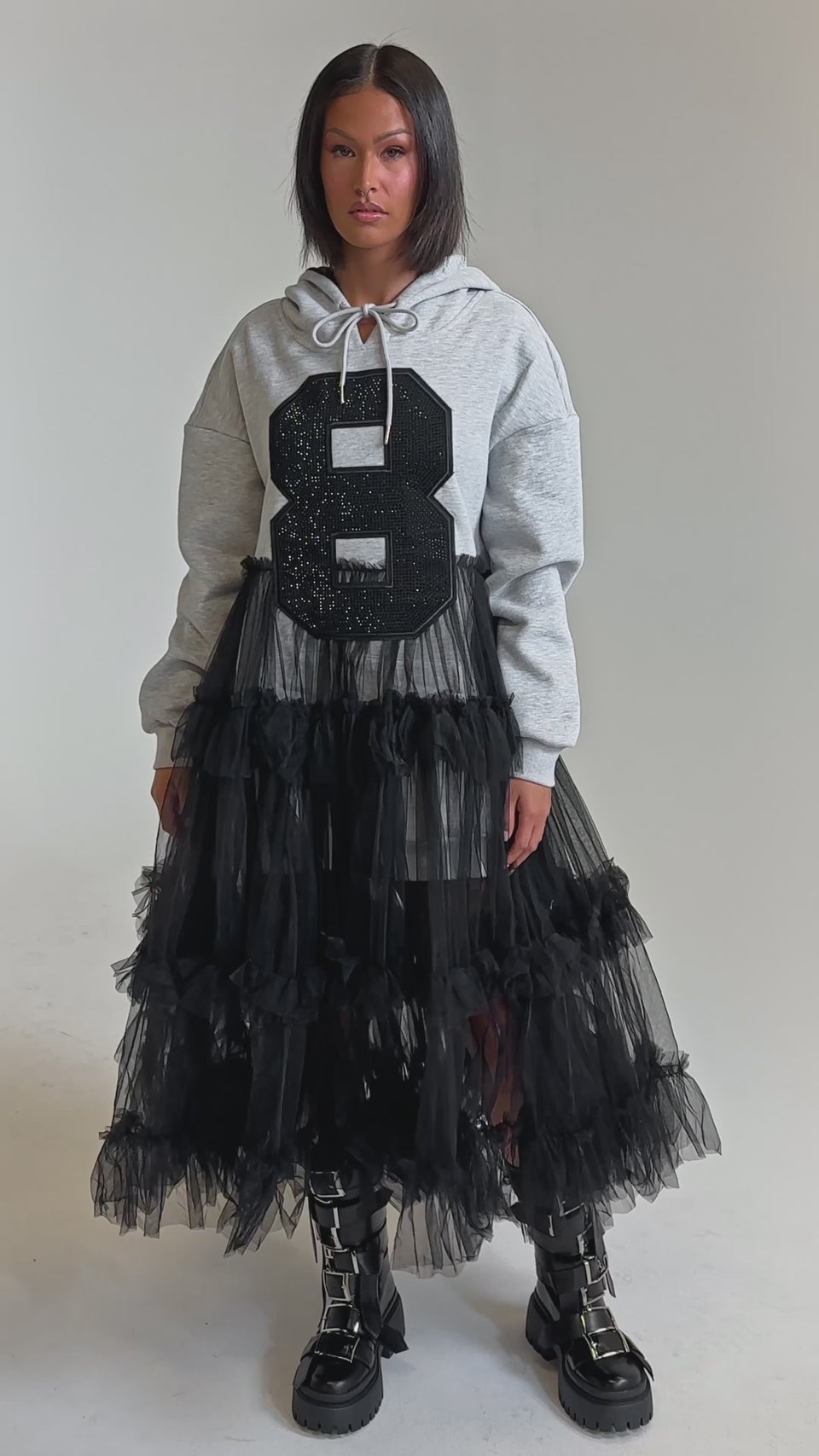 A video of a woman wearing the DO SOMETHING CRAZY SWEATSHIRT DRESS, a gray rhinestone varsity-style hoodie dress with a bold black "8" on the front, styled with a black tulle ruffle maxi skirt and chunky combat boots against a light background.