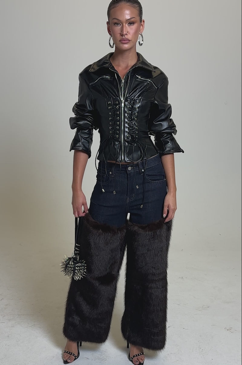 A video of a woman in BOOTS WITH THE FUR WIDE LEG DENIM—wide leg denim pants with brown faux fur trim—paired with a black leather corset jacket, black heels, and a studded mini handbag against a plain gray background.